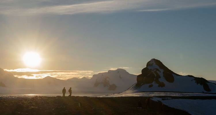 Two figures stand on a snowy headland admiring a low sun over jagged white peaks and a shimmering bay.