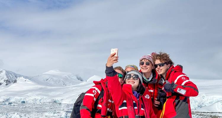 Five bundled friends in matching red parkas snap a cheerful selfie against a backdrop of icy mountains and clouds.