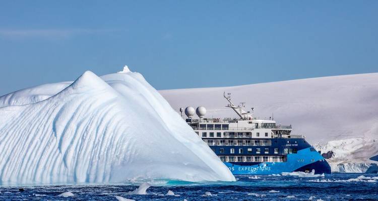 A blue expedition cruise ship sails past a giant sculpted iceberg amid brilliant blue seas and snow-covered hills.
