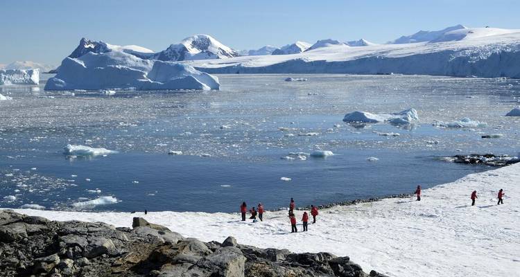 Bright-coated visitors explore a snowy shoreline overlooking a vast iceberg-filled bay under clear blue skies.
