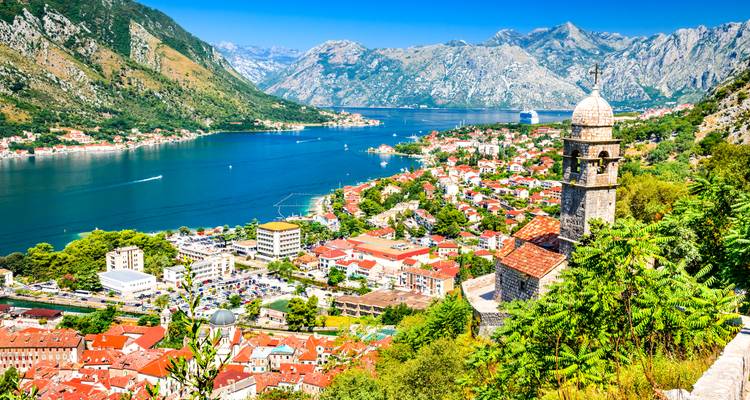 Vista panorámica sobre la Bahía de Kotor con el pueblo histórico, campanario de piedra y montañas empinadas reflejadas en aguas azul profundo.