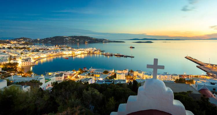 Vue du coucher de soleil sur la ville de Mykonos et son port, vue depuis une chapelle blanchie à la chaux surmontée d'une croix.