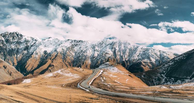 Montañas del Cáucaso cubiertas de nieve y carretera serpenteante bajo nubes dramáticas y luz solar.