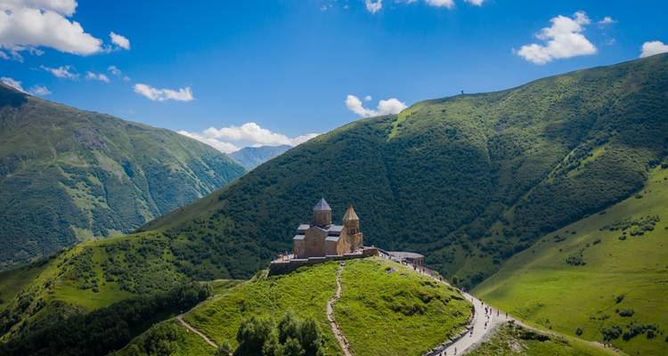 Vista aérea amplia de la Iglesia de la Trinidad de Gergeti aislada en una ladera montañosa verdosa bajo un cielo azul brillante.