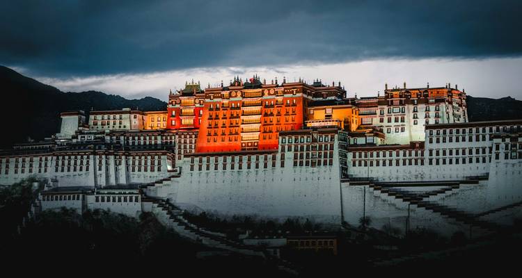 The Potala Palace glows in warm light beneath a dramatic dark sky over the Tibetan plateau.