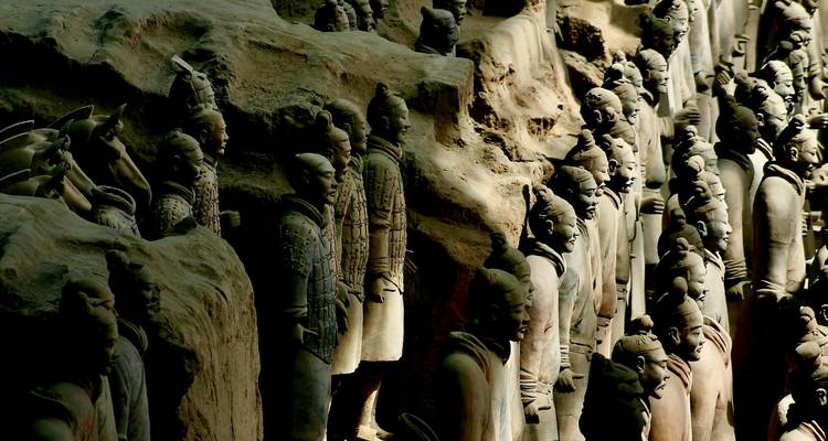Rows of Terracotta Warriors stand partially illuminated inside an archaeological excavation.