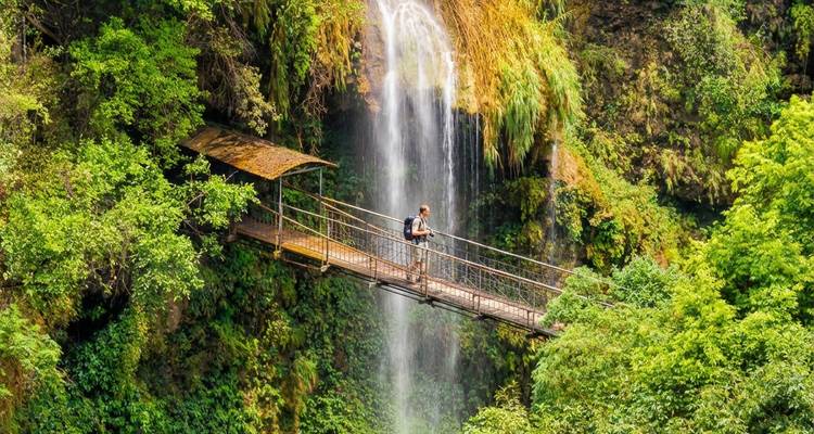 Un routard traverse un pont suspendu étroit devant une cascade dans la jungle.
