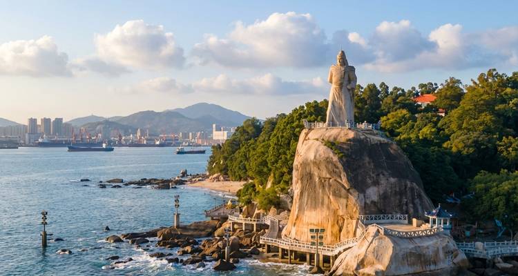 Falaise côtière couronnée par une imposante statue de pierre surplombant un port animé et des montagnes.