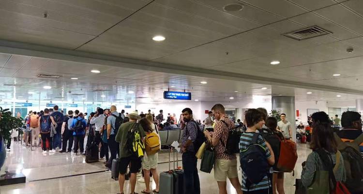 Travellers with suitcases wait in a snaking line inside a bright arrivals hall.
