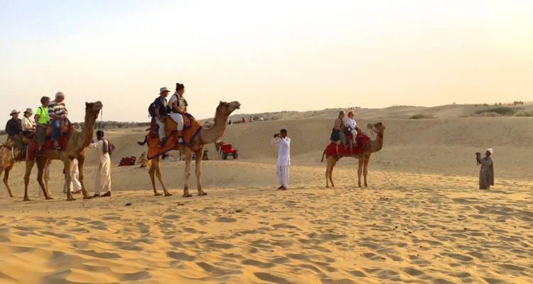 Des touristes chevauchant des chameaux en file indienne à travers les dunes dorées du désert sous un ciel brumeux du soir.