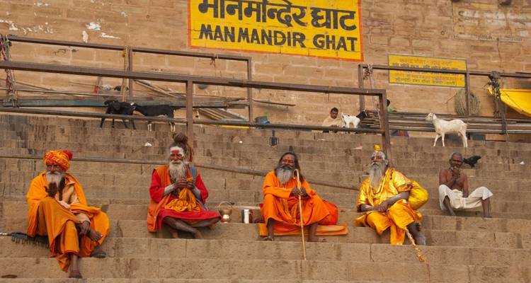 Vier kleurrijk geklede sadhu's zitten op stenen trappen onder een bord bij Man Mandir Ghat in Varanasi.