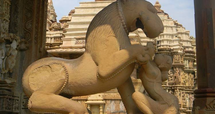 Close-up van een zandstenen leeuwsculptuur die een figuur aanvalt tegen de sierlijke achtergrond van Khajuraho tempels.