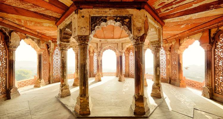 Zonlicht stroomt door bogen en gebeeldhouwde zuilen van een open Mogolpaviljoen met een gepatroneerd plafond.