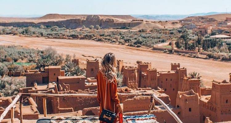 Woman in red dress looks over the earthen kasbah buildings of Ait Benhaddou with a riverbed below.