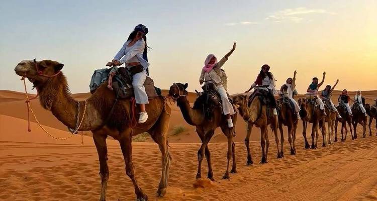 Line of riders atop camels waving as they trek across orange sand at sunset.