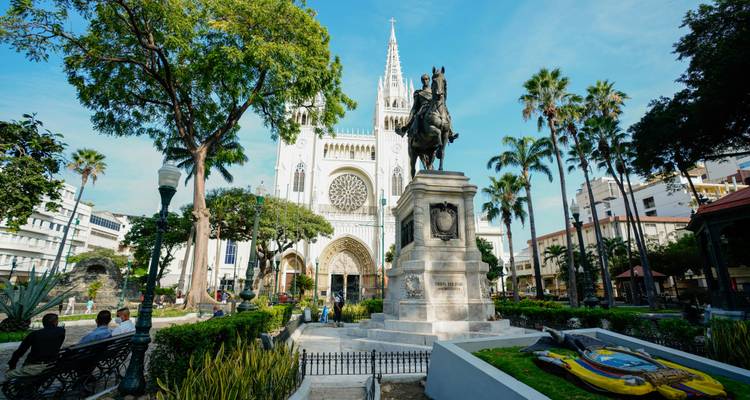 Plaza rodeada de estatuas con catedral neogótica, árboles frondosos y gente local relajándose en los bancos.
