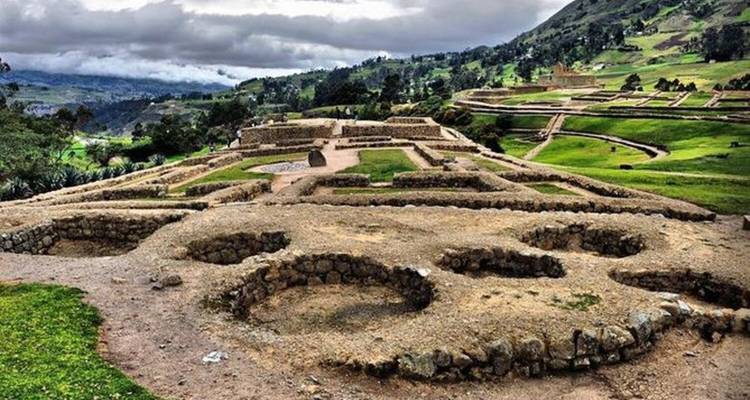 Ruinas de piedra con cimientos circulares se encuentran entre verdes colinas ondulantes y nubes dramáticas en las tierras altas andinas.