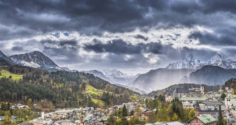 Un dramático pueblo alpino se acurruca en un valle verde bajo picos nevados y un cielo tormentoso con rayos de luz.