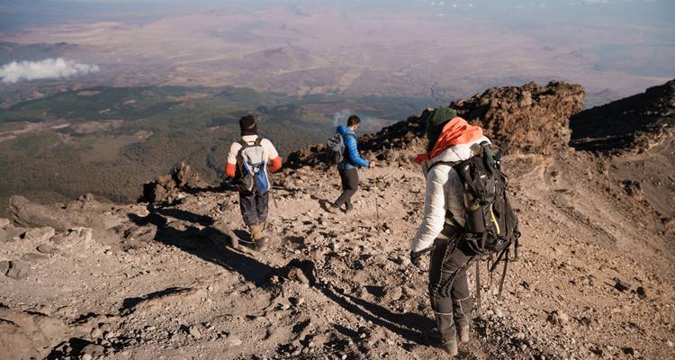 Tres excursionistas descienden cuidadosamente por una empinada ladera volcánica polvorienta en las alturas del Kilimanjaro con amplias vistas del valle abajo