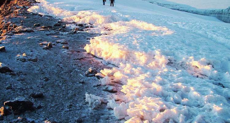 Dos pequeños escaladores caminan a lo largo de una cresta de hielo dramática que brilla rosa bajo el sol matutino en las alturas del Kilimanjaro