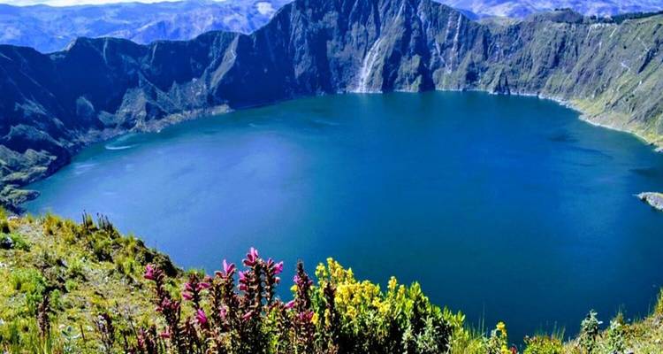 Vivid wildflowers frame the turquoise crater lake of Quilotoa with steep volcanic walls under clear skies.
