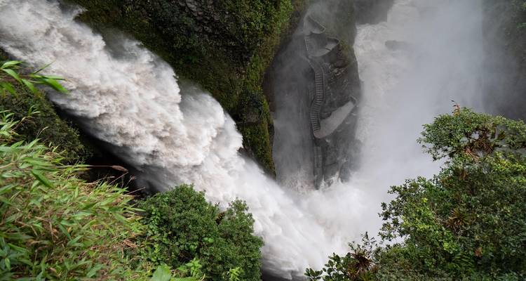 Aerial view of a roaring waterfall plunging through lush greenery with a winding staircase carved into rock.