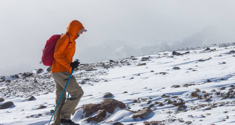 Excursionista solitario con chaqueta naranja usando bastones de trekking contra un telón de fondo alpino nevado y azotado por el viento