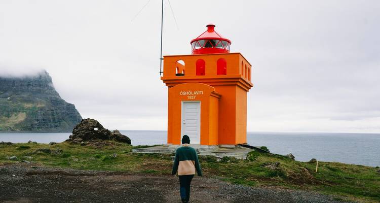 Une personne portant un bonnet marche vers un phare orange éclatant perché sur une falaise herbeuse surplombant la mer sous un ciel nuageux.