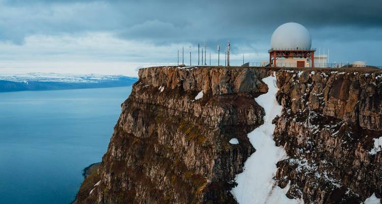 Station radar au sommet d'une falaise spectaculaire aux traînées de neige surplombant un vaste fjord bleu.