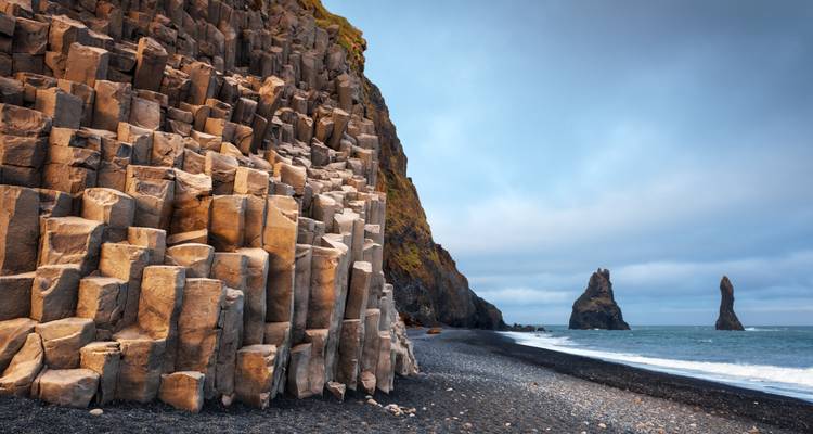 Des colonnes de basalte impressionnantes bordent une plage de sable noir avec les formations rocheuses de Reynisdrangar visibles sous un ciel menaçant.