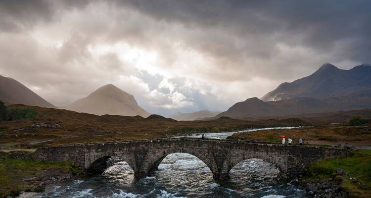 Stenen boogbrug over een bruisende rivier met dramatische bergen en stormachtige luchten op het eiland Skye
