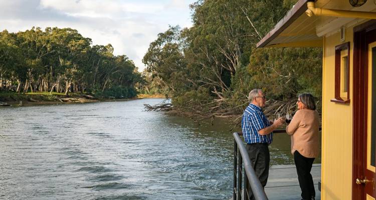 Una pareja mayor disfruta de bebidas en la cubierta de una casa flotante navegando por un río ancho bordeado de eucaliptos.