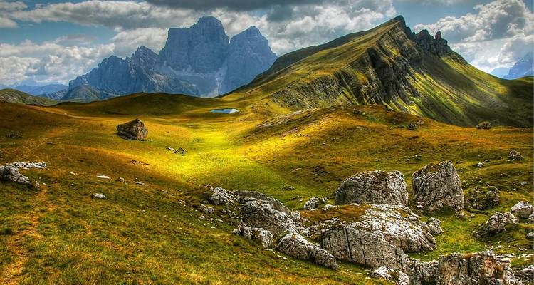 Plateau alpin ondulant et ensoleillé avec des pics rocheux spectaculaires et une herbe d'un vert éclatant sous des nuages tourmentés.