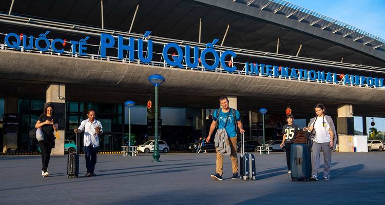 Travelers pull suitcases while walking away from the Phu Quoc airport terminal under clear blue skies.