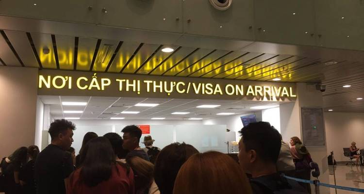 Passengers queue beneath a bright yellow "Visa on Arrival" sign inside an airport immigration area.