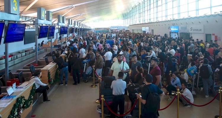 A packed check-in hall with long lines of passengers and luggage inside a bright airport terminal.