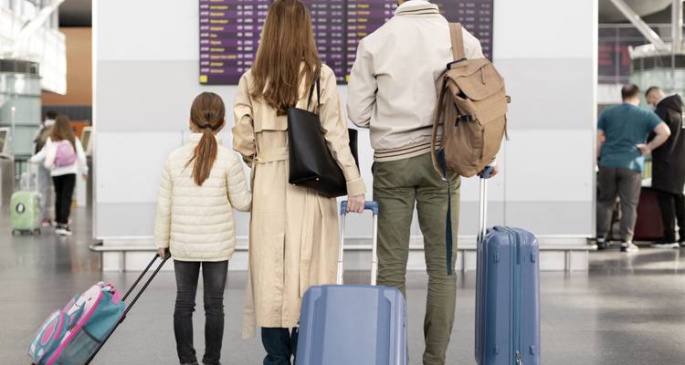 Rückansicht einer Familie mit Koffern, die auf eine Abflugstafel am Flughafen schaut.