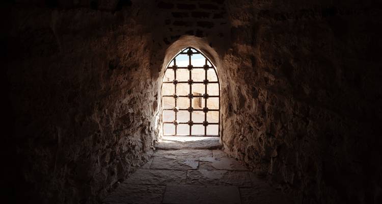 Pasillo de piedra tenuemente iluminado que conduce a una pequeña ventana arqueada con barras de hierro y luz del día más allá.