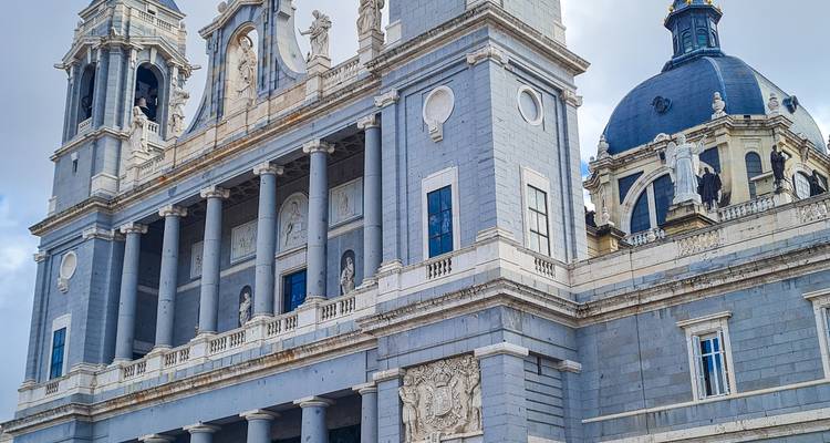 Steingraue Türme und Kuppeln der Almudena-Kathedrale in Madrid vor bewölktem Himmel.