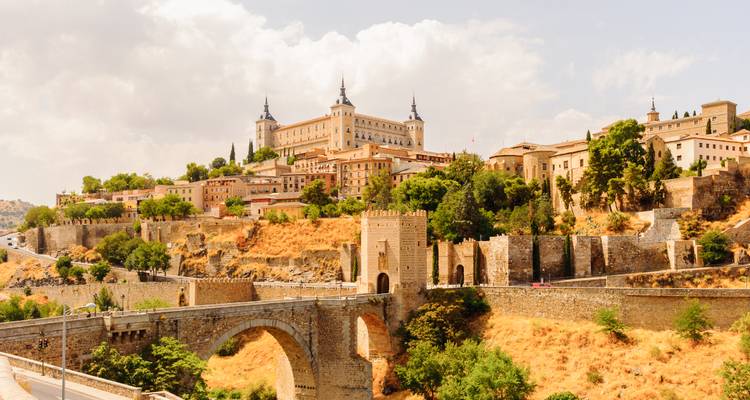 Vista panorámica del Alcázar de Toledo dominando una ciudad histórica en la cima de una colina con un puente de piedra cruzando un río abajo en un día soleado y brillante
