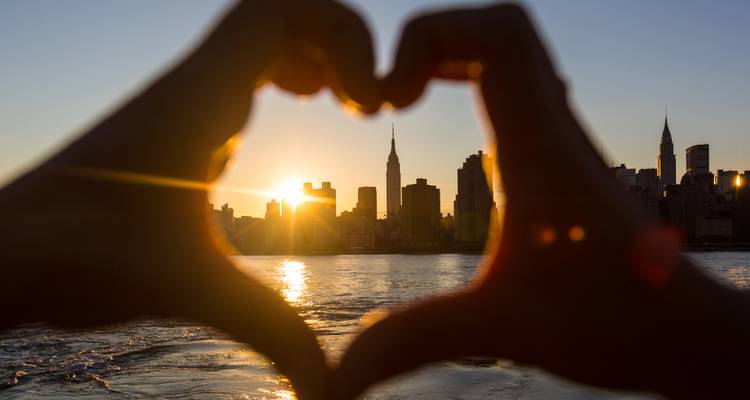 Silueta en forma de corazón formada por las manos enmarca el horizonte de Manhattan y el sol poniente al otro lado del río.