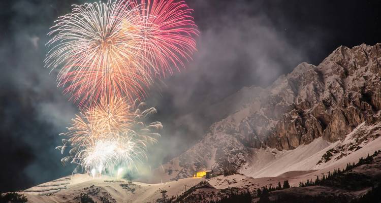Bunte Feuerwerke explodieren über verschneiten Alpenhängen und schroffen Gipfeln unter einem sternenklaren Nachthimmel.