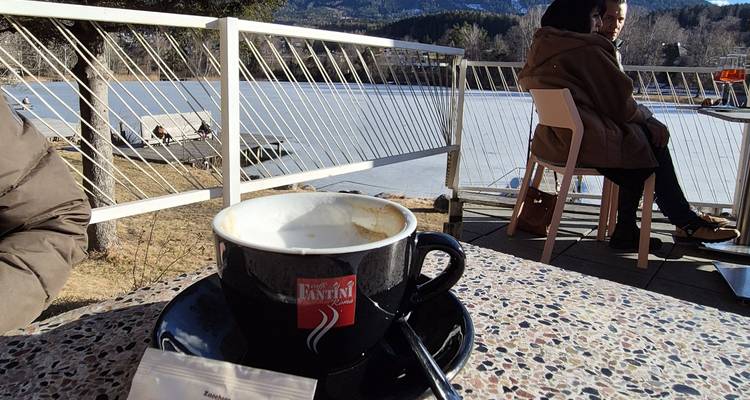 Ein halb ausgetrunkener Cappuccino steht auf einem Terrassentisch mit Blick auf einen gefrorenen See und entfernte Hügel, während Cafébesucher in der Nähe plaudern.