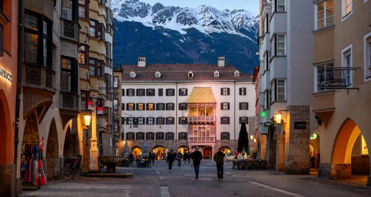 Abendlichter leuchten in einer schmalen Fußgängerstraße von Innsbruck, eingerahmt von bunten Fassaden und schneebedeckten Gipfeln in der Ferne.