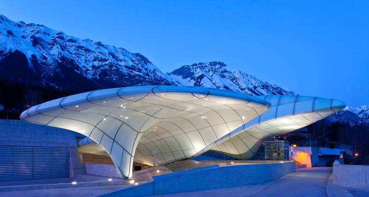 Die futuristische Glas-und-Stahl-Hungerburgbahn-Station leuchtet bei Abenddämmerung blau vor den Alpengipfeln.