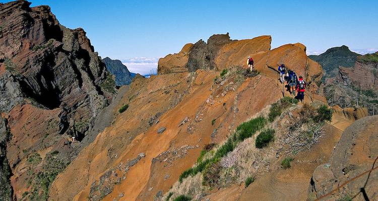 Wandelaars trekken langs een dramatische oranje-rode rotsrichel met steile kliffen en een heldere blauwe lucht op de achtergrond.