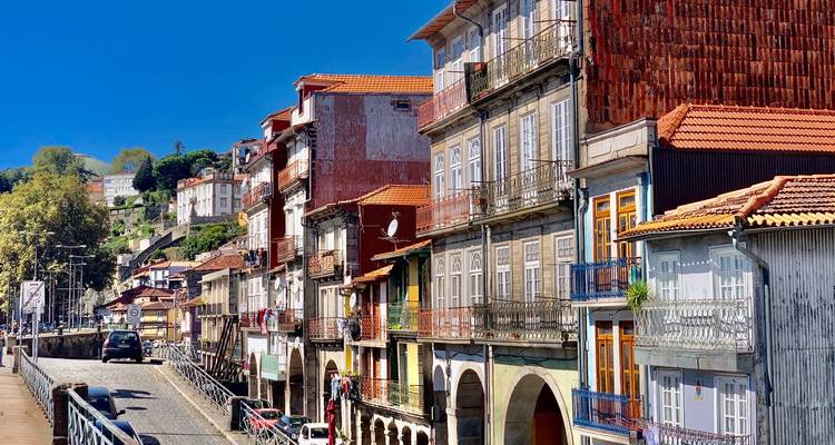 Bunte Stadthäuser am Flussufer säumen eine gepflasterte Straße unter einem klaren blauen Himmel.