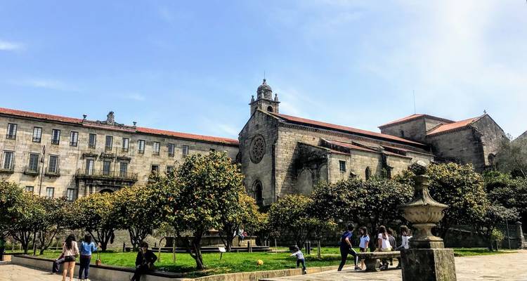 Besucher entspannen sich auf einem grünen Platz neben einem historischen Steinkloster unter blauem Himmel.