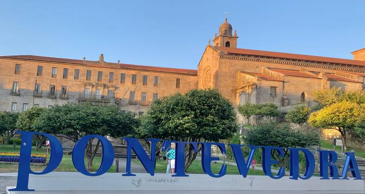 Große blaue Buchstaben buchstabieren PONTEVEDRA vor einem eleganten historischen Gebäude im Abendlicht.