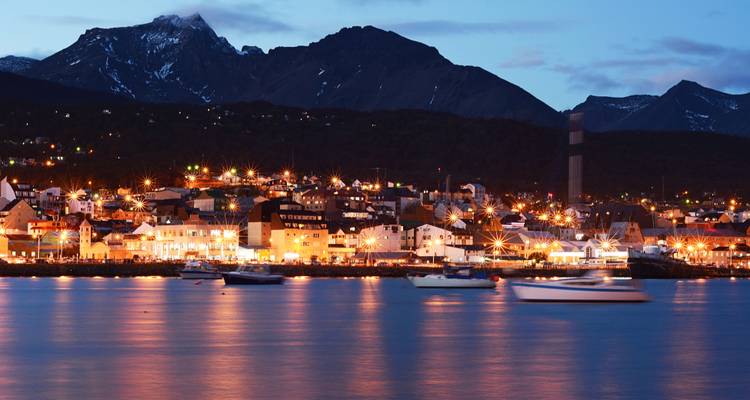 Lumières du soir d'Ushuaia reflétées sur la baie calme avec les montagnes sombres derrière.